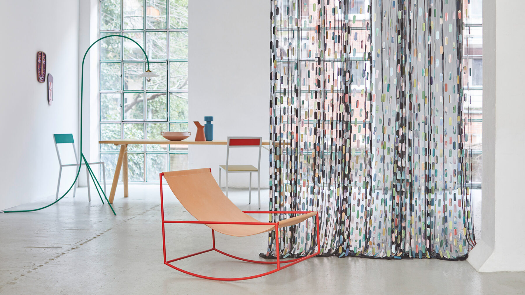 ein Schaukelstuhl mit rotem Metallrahmen und Sitzfläche aus Leder vor einem transparenten Vorhang mit Pailletten, im Hintergrund ein Tisch mit Stühlen und eine Stehlampe vor großen Fensterfronten