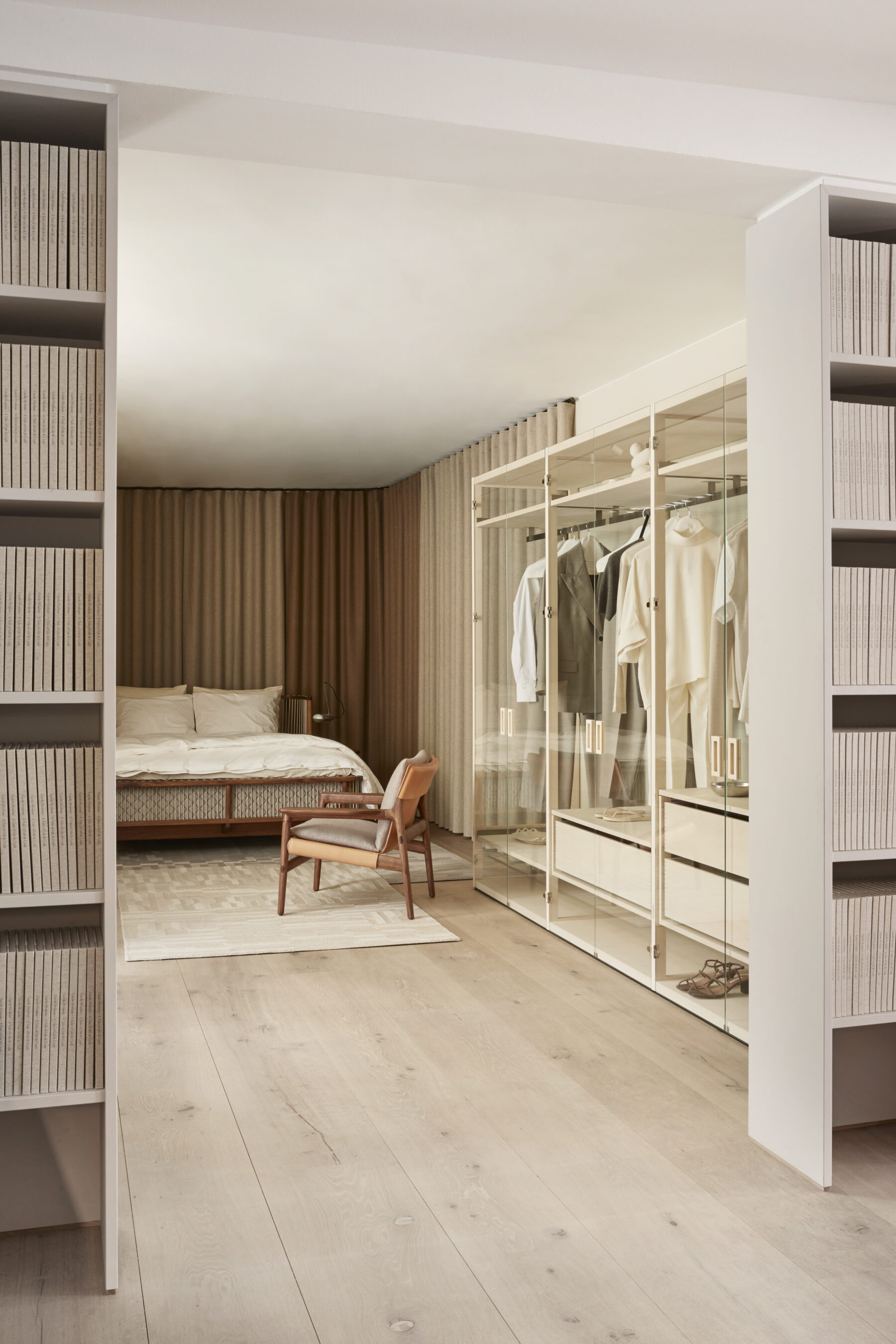 Von zwei Bücherregalen gesäumtes Schlafzimmer Interieur von Garde Hvalsøe mit einem Schranksystem aus Holz mit gläsernen Fronten auf der rechten Seite, davor ein Holzsessel, im Hintergrund ein elegantes Doppelbett aus Holz.